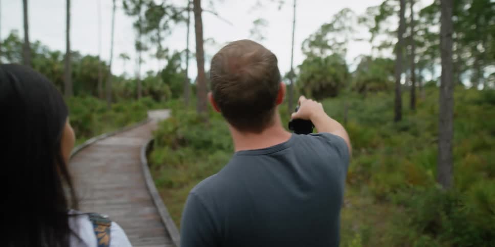 Man holds a camera and points to the trees as he walks on a trail with a woman