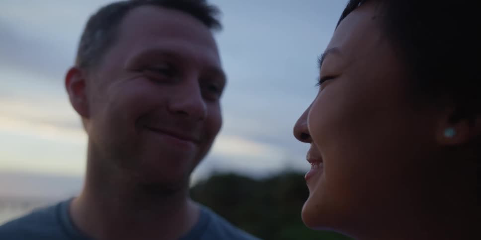 Man and woman look lovingly at each other while standing near a lake with a forest in the distance