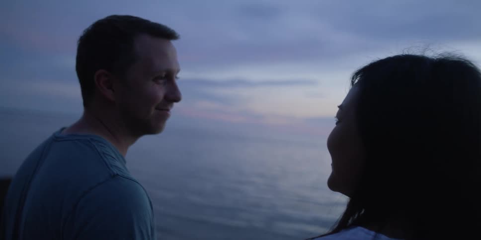 A man and woman smile at each other while looking at the ocean at sunset