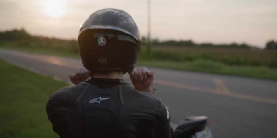 A man in riding jacket puts on his helmet as he approaches and climbs onto his motorcycle parked beside a road in the countryside on a cloudy evening