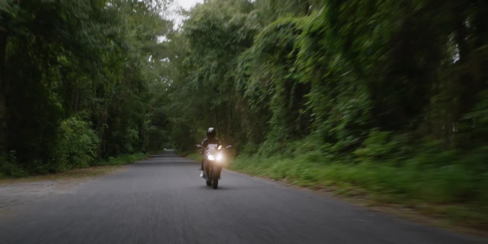 Cyclist wearing a black jacket and black helmet is riding a motorcycle with the headlights on down a road surrounded by trees