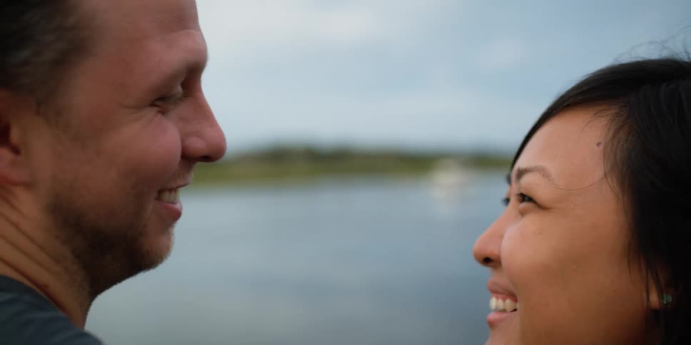 A couple smile at each other as they stand near a lake