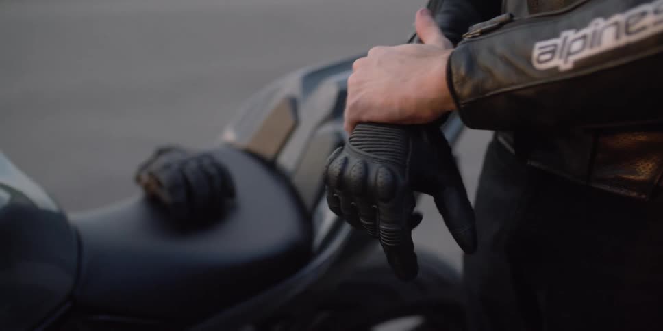 A person with a leather jacket putting on gloves with a motorcycle and a road in the background