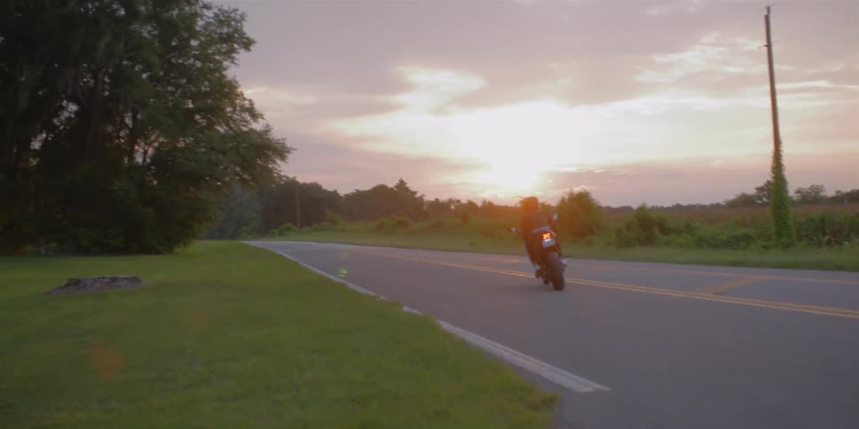 A person wearing a crash helmet rides a motorcycle on a rural road at sunset