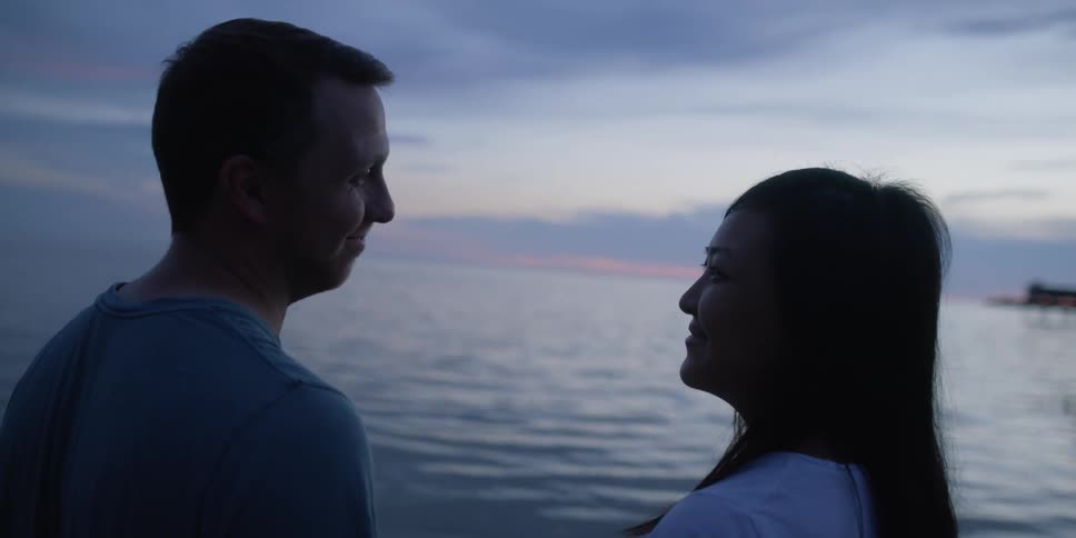 A man and a woman standing by a lake with the sky and clouds in the background