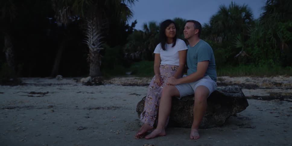 A couple sit on a large rock on the beach holding hands and looking toward the ocean