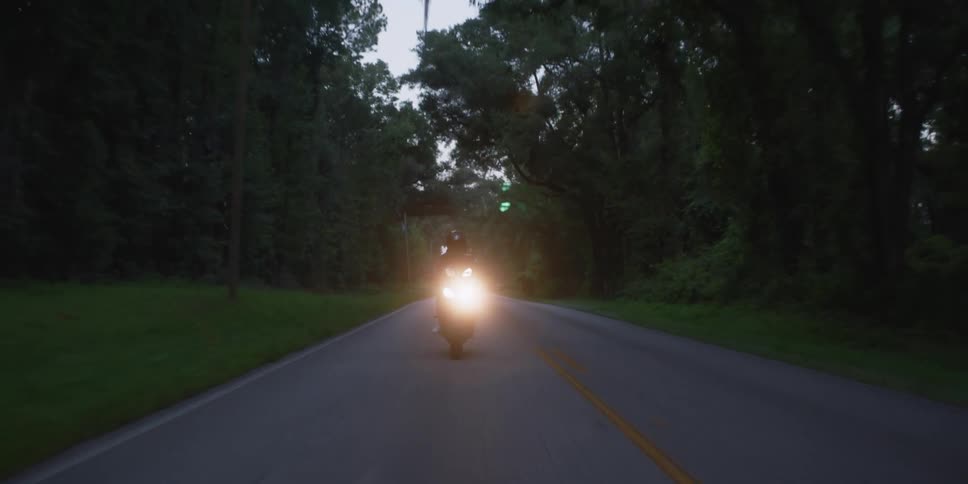 Cyclist wearing a black jacket and black helmet is riding a motorcycle with the headlight on down a road surrounded by trees