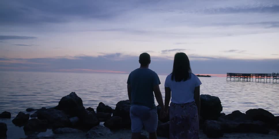 A man holds his girlfriends hand as he leads her toward rocks along the shore of a lake at sunset