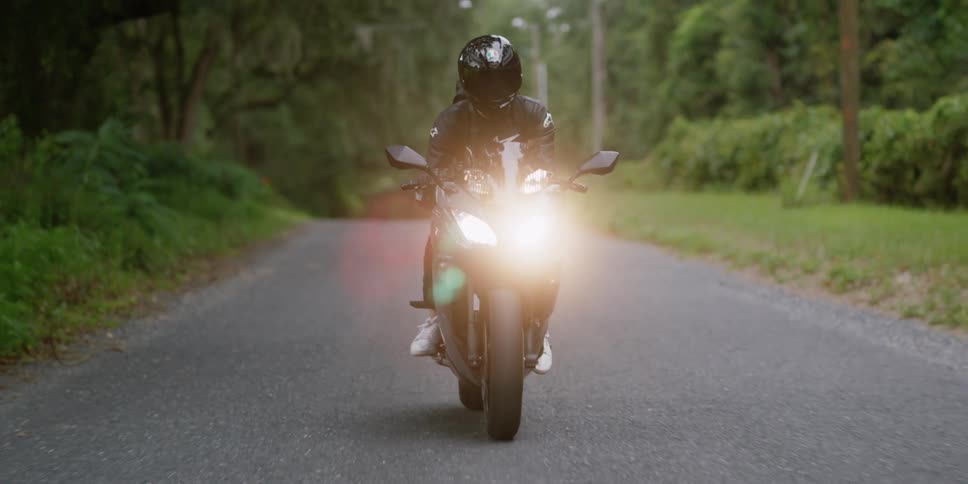 A person with a helmet riding a motorcycle on a road surrounded by trees with the sky and clouds in the background