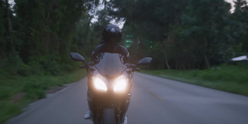 A person with a helmet riding a motorcycle on a road surrounded by trees with the sky and clouds in the background