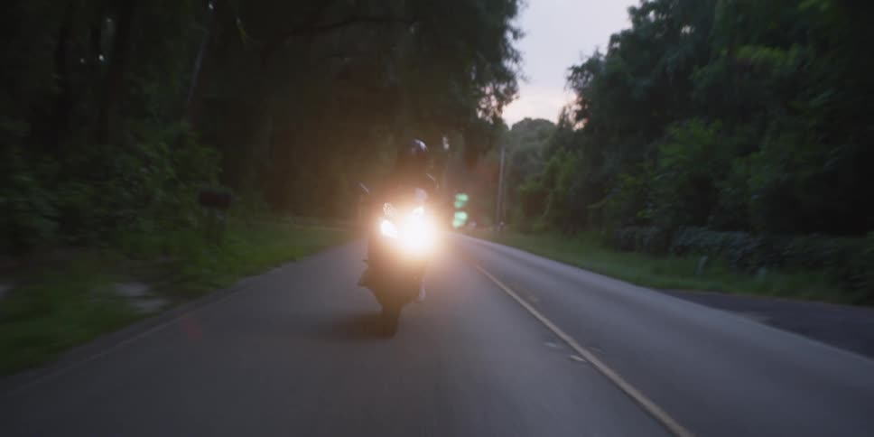 Cyclist wearing a black jacket and black helmet is riding a motorcycle with the headlights on down a road surrounded by trees