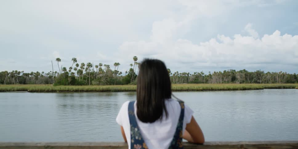 A woman on a dock by a lake fishing with a fishing pole with trees the sky and clouds in the background