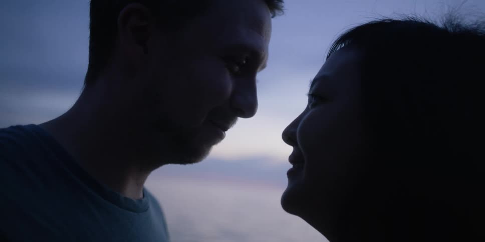 A man and a woman standing by a lake with the sky and clouds in the background