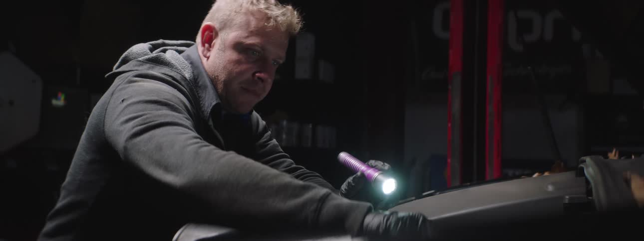 A man in a garage with a flashlight and gloves working on a vehicle