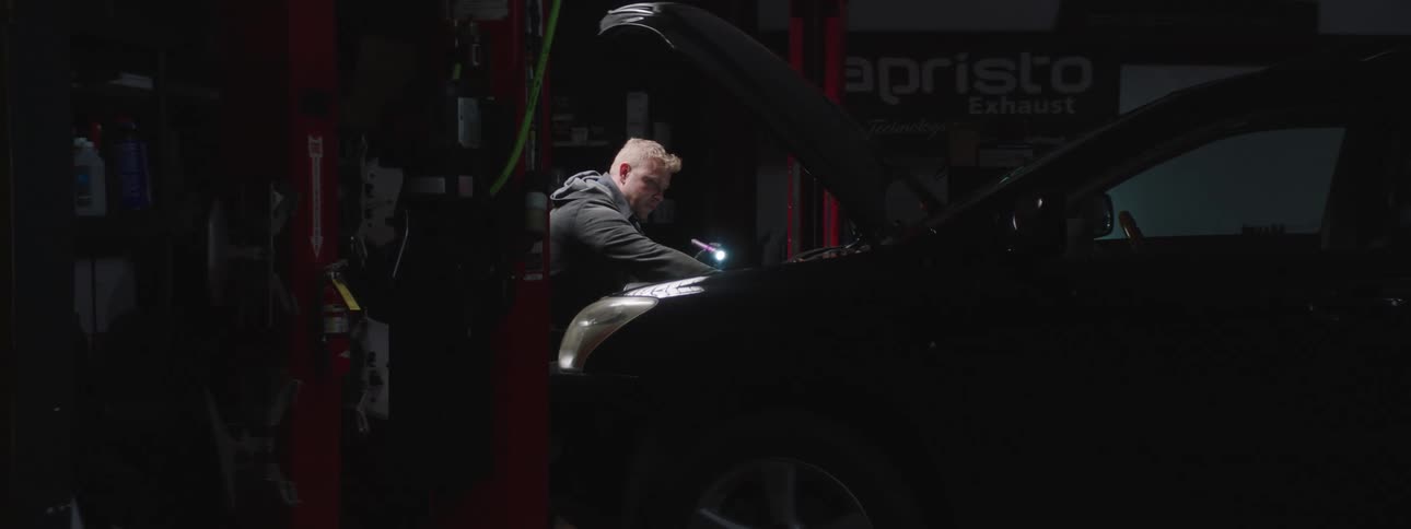 A male auto mechanic stands in a darkened garage while inspecting an auto engine