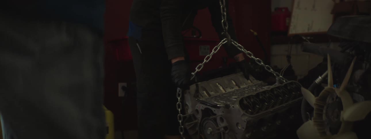 A mechanic repeatedly pushes a lever to control a hoist with which he is lifting an auto engine