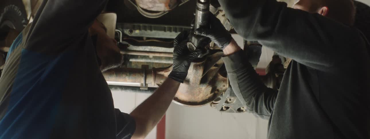 Two mechanics inspect components on the underside of a car on a car lift above them