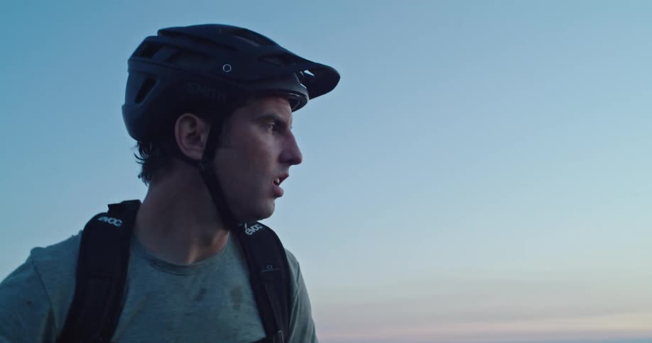 A man breathes heavy resting his arms on handlebars then stands and look at hills and sky at sunset