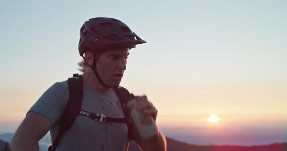 A man reaches the summit of a mountain on his bike and takes a drink of water at sunset