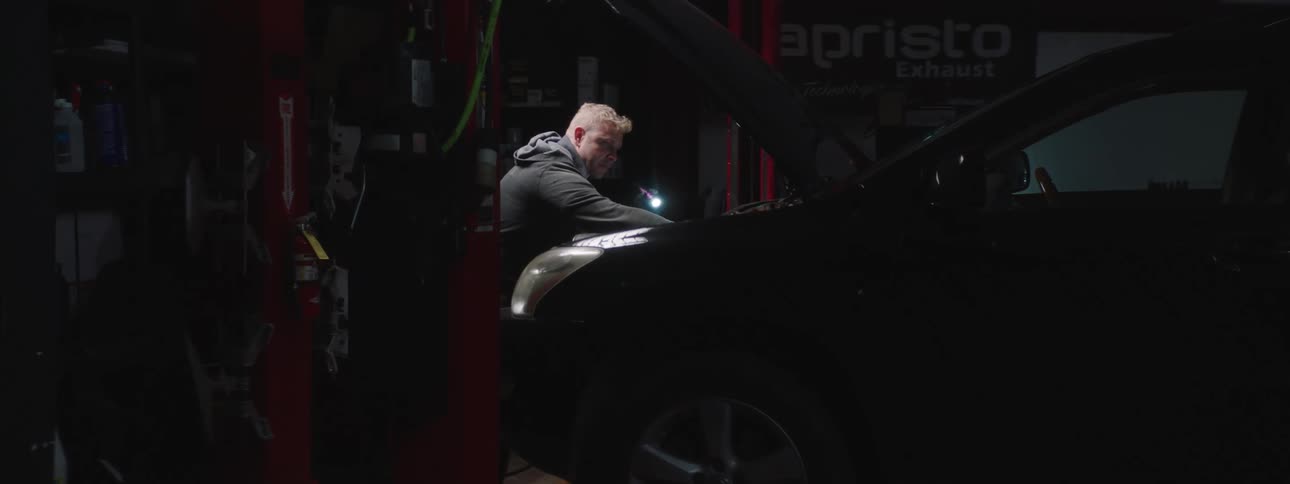 A man in a garage with a flashlight working on a vehicle with a wall in the background