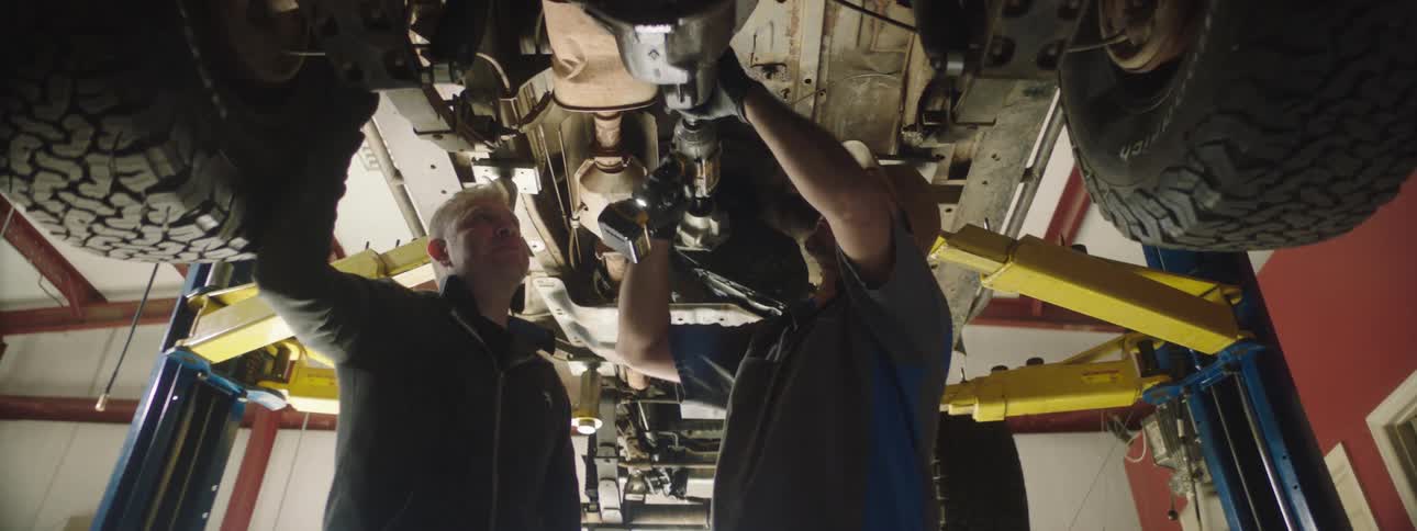 Two men in a garage working on a vehicle with a wall and window in the background