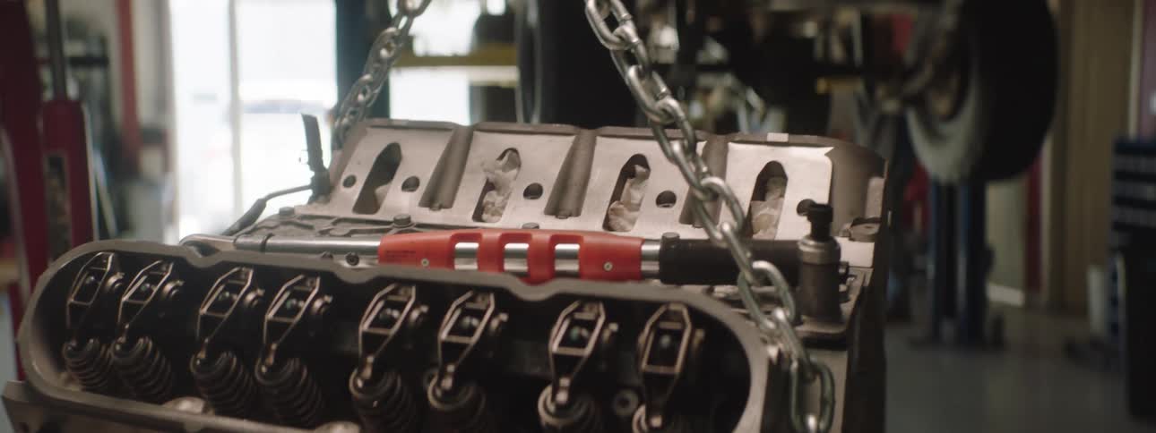 Engine hangs from a chain near a truck that is raised on a truck lift in a garage while two people stand near it