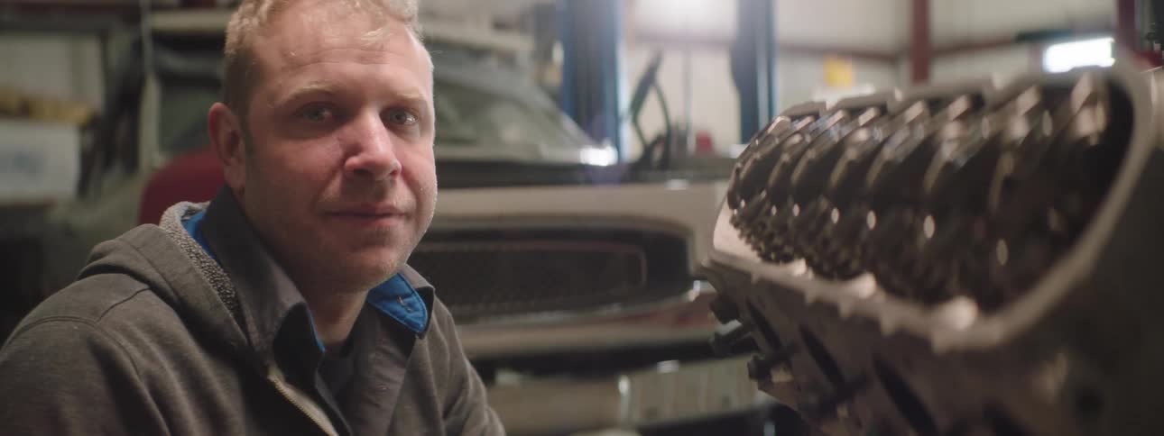 Blonde car mechanic with grey shirt and jacket looks at the camera smiling at the garage
