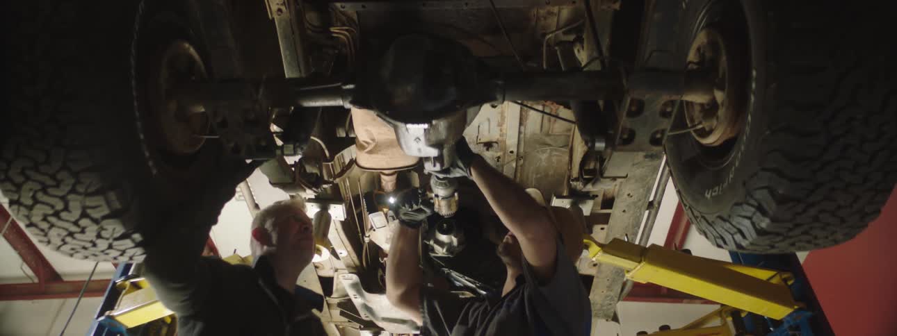 Mechanic uses a tool on the underside of a vehicle that is raised on a car lift while another man watches