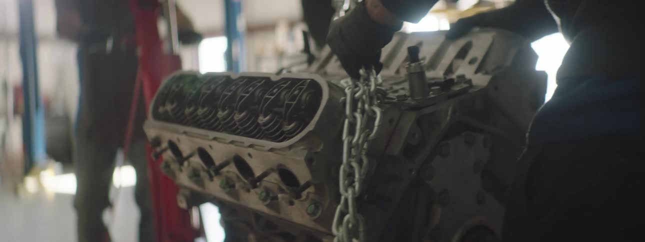 Two men in the garage moving an engine with a vehicle and a wall in the background