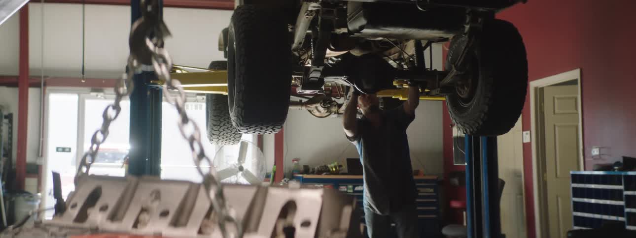 Man works on the underside of a truck that is on a lift as another man approaches him