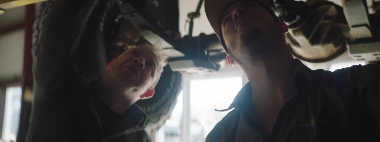 Car mechanic with a cap and man stand under the car and pointing at car part to check it at the garage