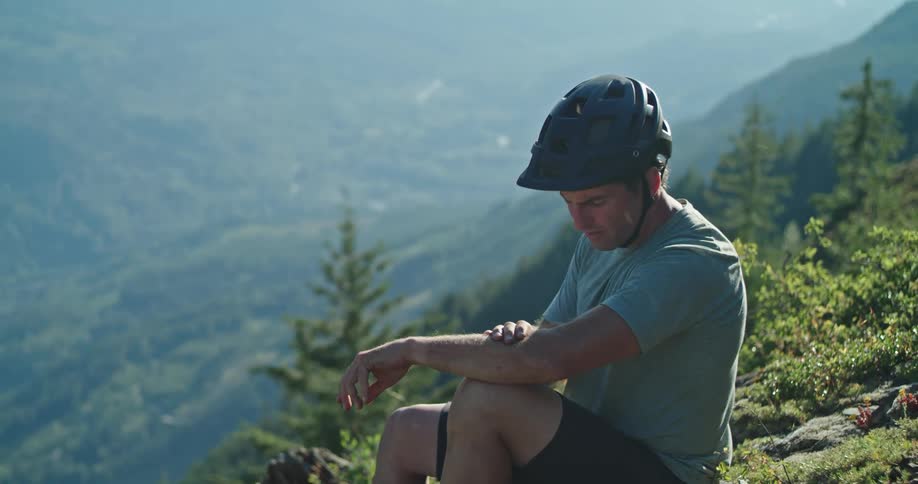 A man sitting outdoors on a mountainside slaps at a bug landing on his arm