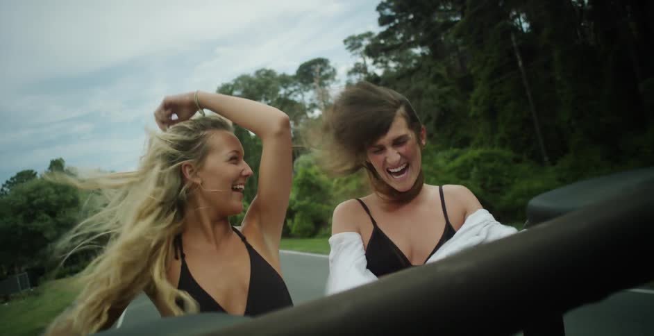 Two young women in bikinis laugh with their hair blowing in the breeze from the back of a vehicle