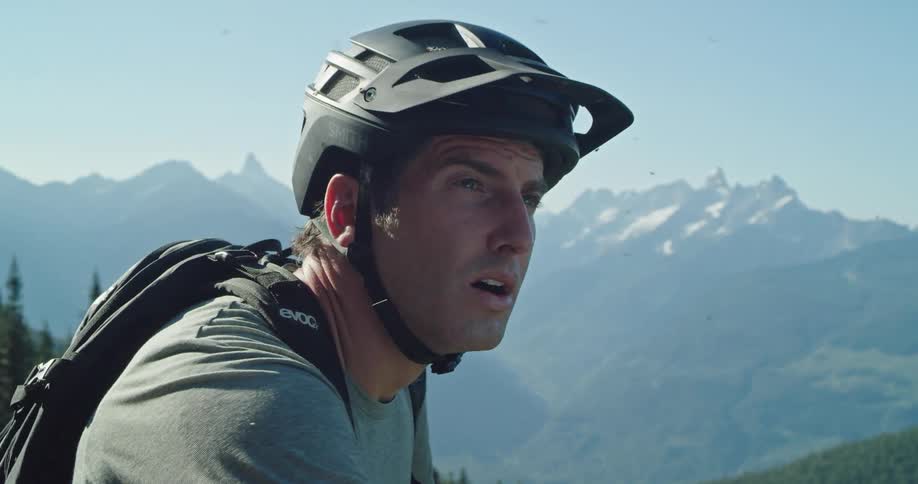 A man with a bicycle helmet and backpack takes a break and looks toward the mountain scenery