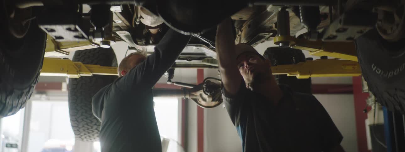Mechanic and customer stand underneath a car on a car lift while the mechanic points to something under the car