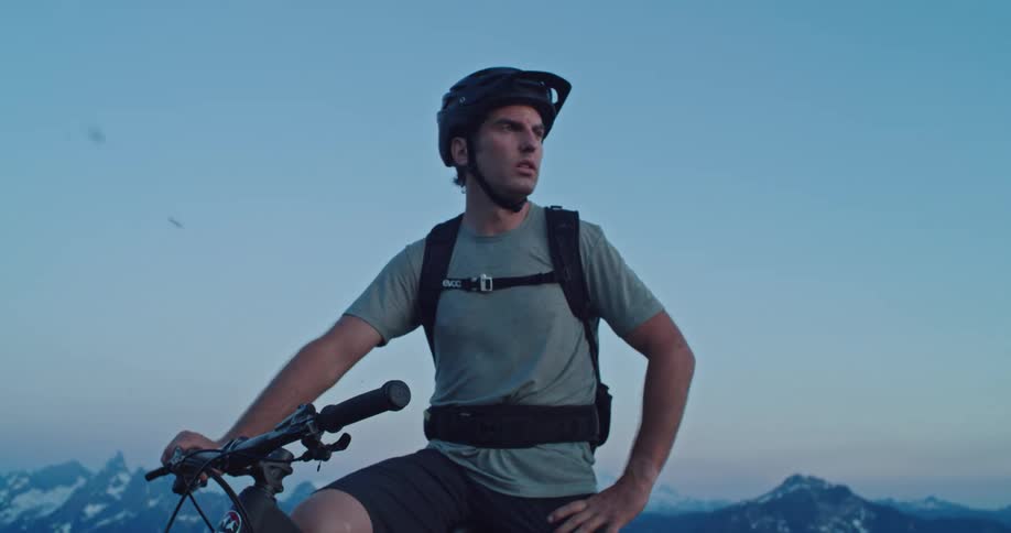 A man rests at the top of a mountain bike summit as the wind blows at dusk