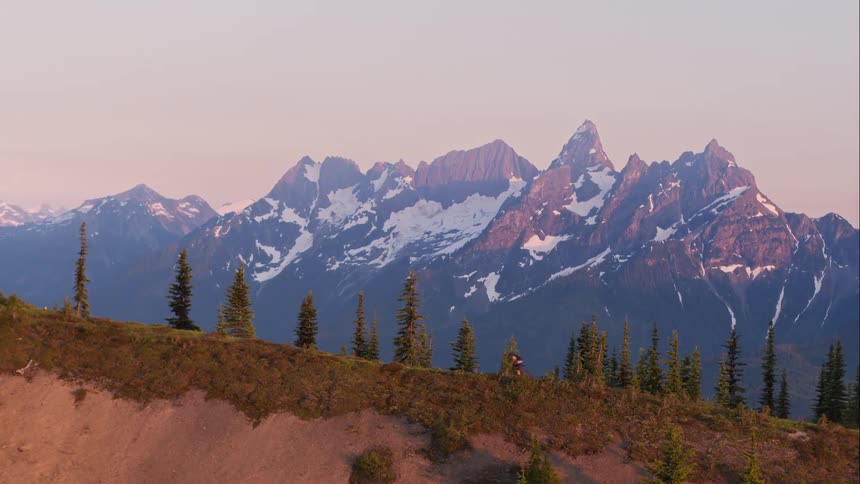 A person on a bicycle rides up a mountain trail with snow covered peaks in the distance