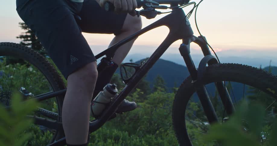 A person sitting on a bicycle and grabbing a water bottle with a mountain range the sky and clouds in the background