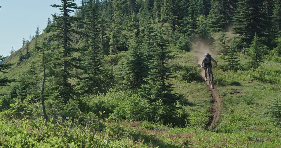 A person with a helmet riding a bicycle on a dirt path surrounded by trees with the sky and clouds in the background
