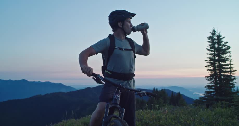 A man pulls a water bottle from his mountain bike and takes drink at dusk on top of a mountain