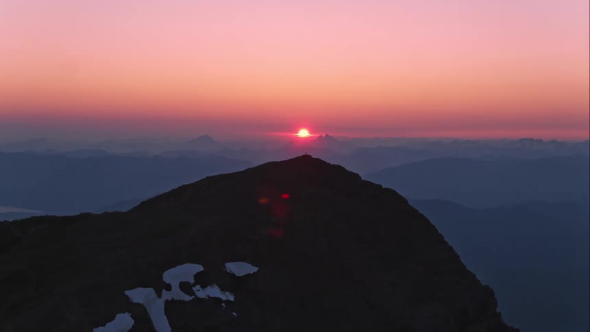 The sun sets in distant horizon as aerial wraps around a mountain top