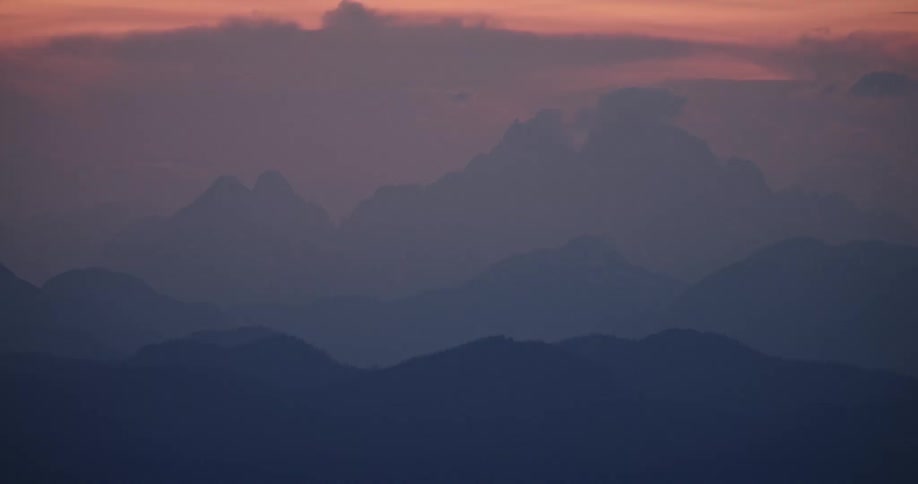 The sun peaks through dense clouds over a jagged distant mountain range
