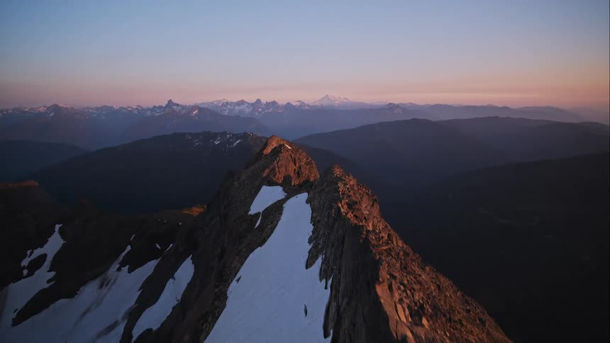 Aerial push over jagged mountain peak at sunset with distant snowy mountain range