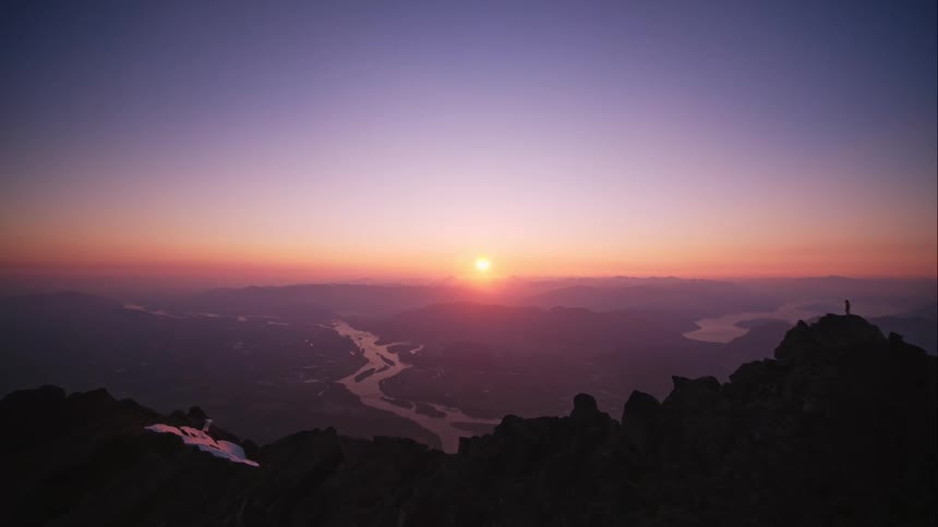 Aerial reveal over mountain peak of river landscape at sunset as person stands on edge of cliff