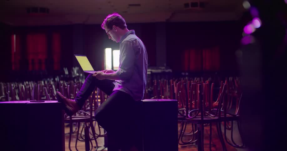 A man leans against a speaker holding a laptop with numerous chairs around him