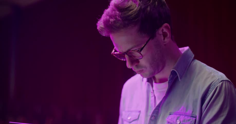 A man types on a laptop resting in his lap while sitting under purple lighting indoors