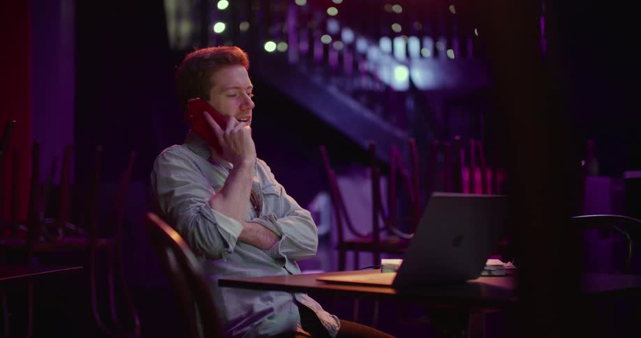 A man sits at a table with a laptop and dials a number on his cell phone