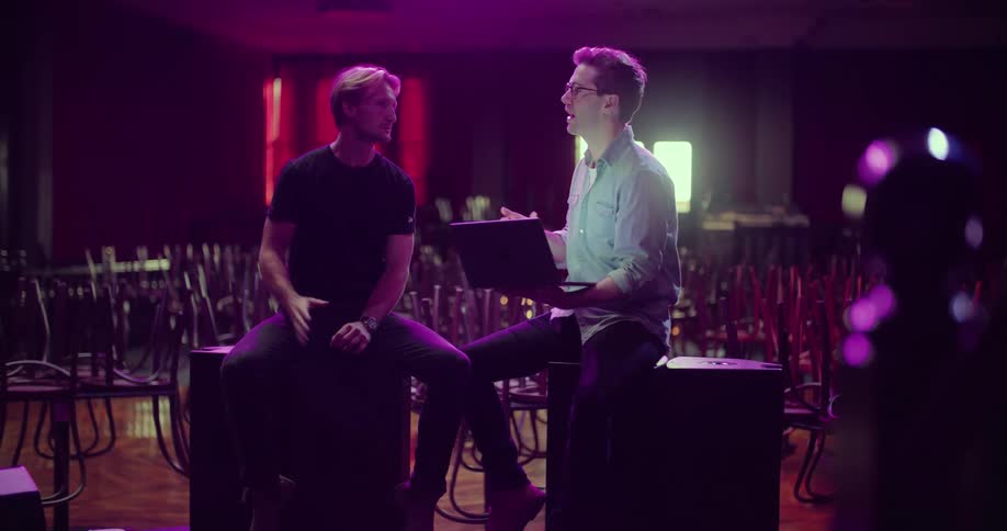 Two men sit on the stage of a small nightclub while discussing some of the layout