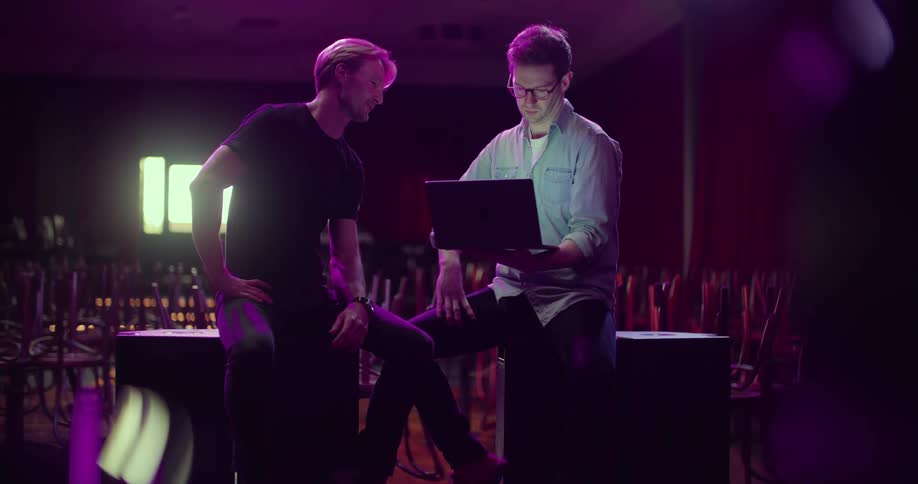 Two men sitting on speakers in a nightclub looking at a laptop with chairs and tables in the background