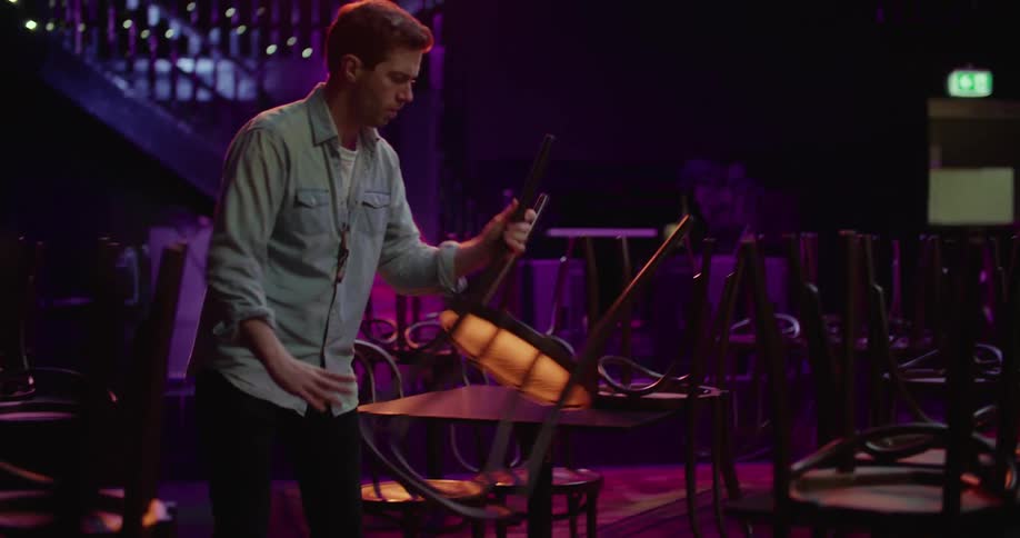 A man in a bar taking chairs from on top of tables and placing them on the floor with a wall and doorway in the background